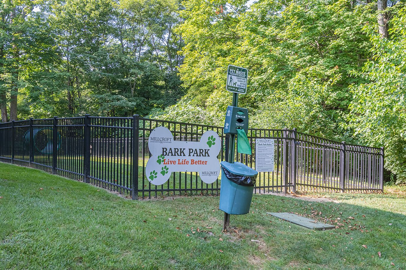 Fenced dog park area with a large Bark Park sign, waste station, and green surroundings.