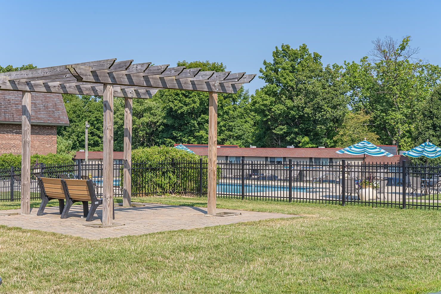 Outdoor community pool area with a wooden pergola, benches, and striped umbrellas behind a black fence.