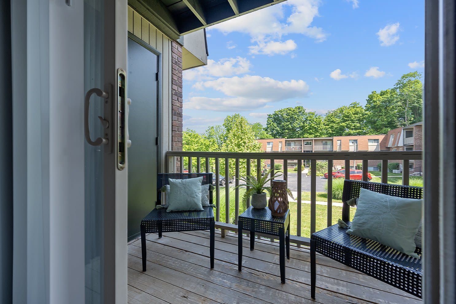 Balcony seating with two black wicker chairs, a small table, cushions, and a plant, overlooking a residential complex.