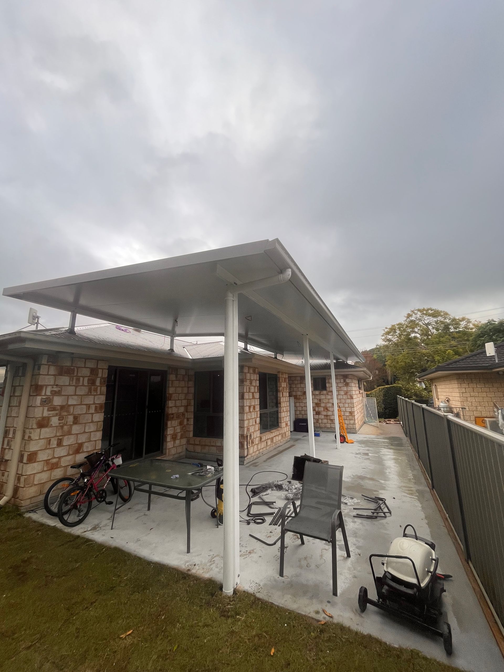Patio with white roof, supported by white posts, with brick house and overcast sky.