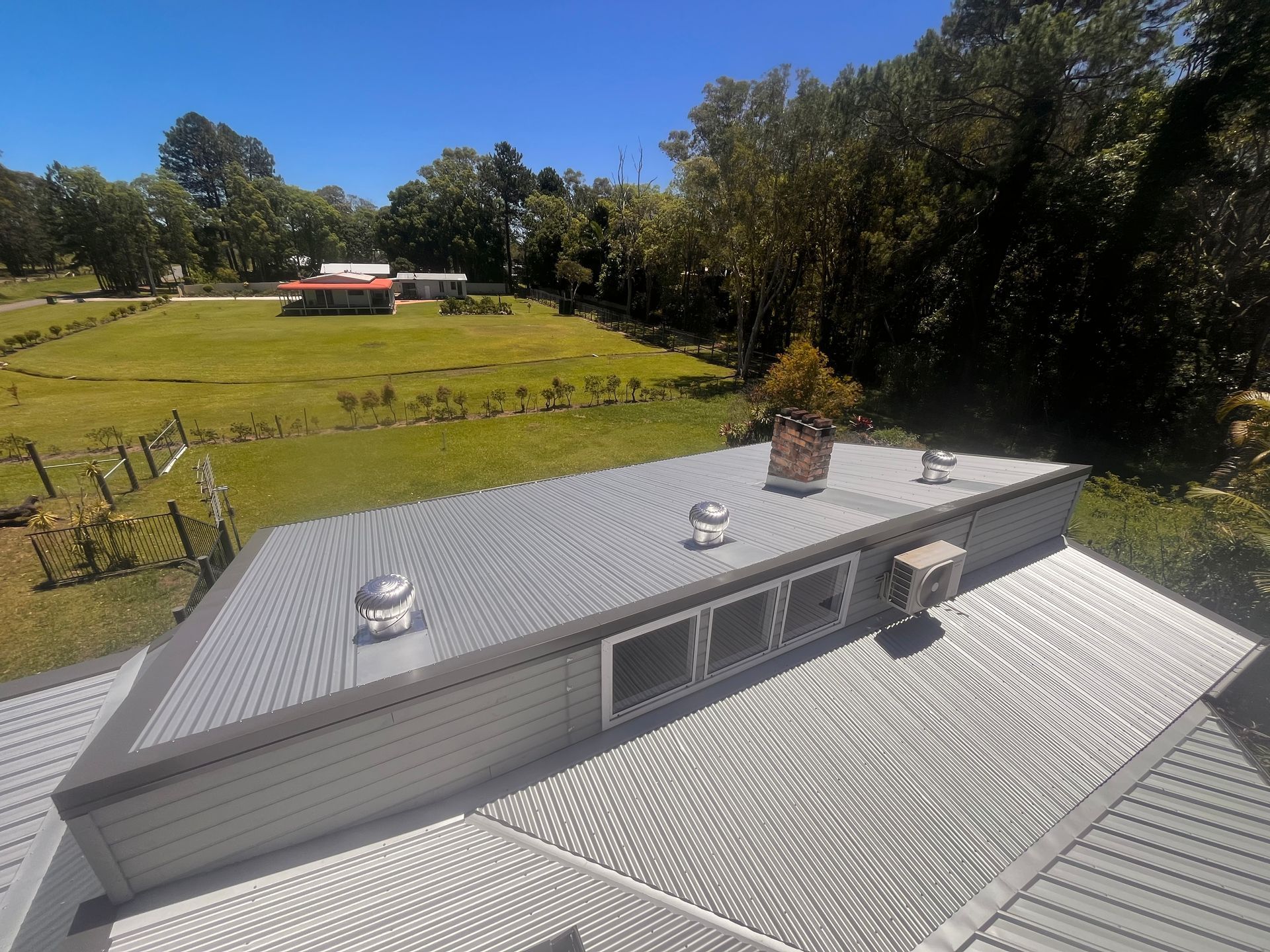 Gray metal roof with vents, overlooking a green field and trees on a sunny day.
