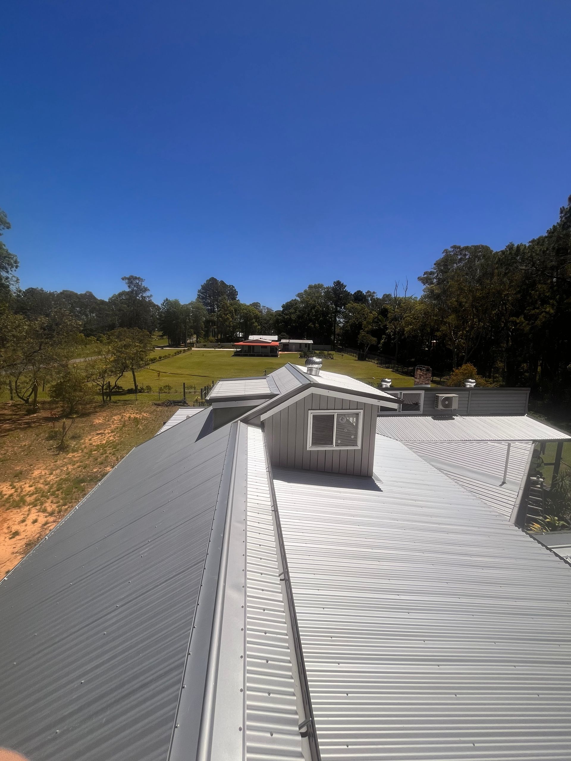 Metal roof of a house with a small structure and a distant red-roofed building under a blue sky.