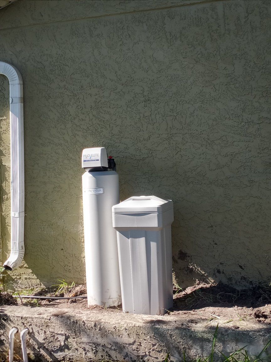 Water softener system against a textured beige wall, with a white drainpipe to the left.