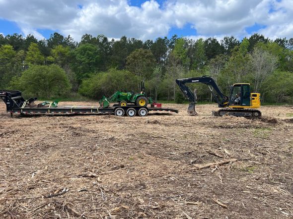 Excavator and trailer with machinery on a cleared wooded area, blue sky in background.