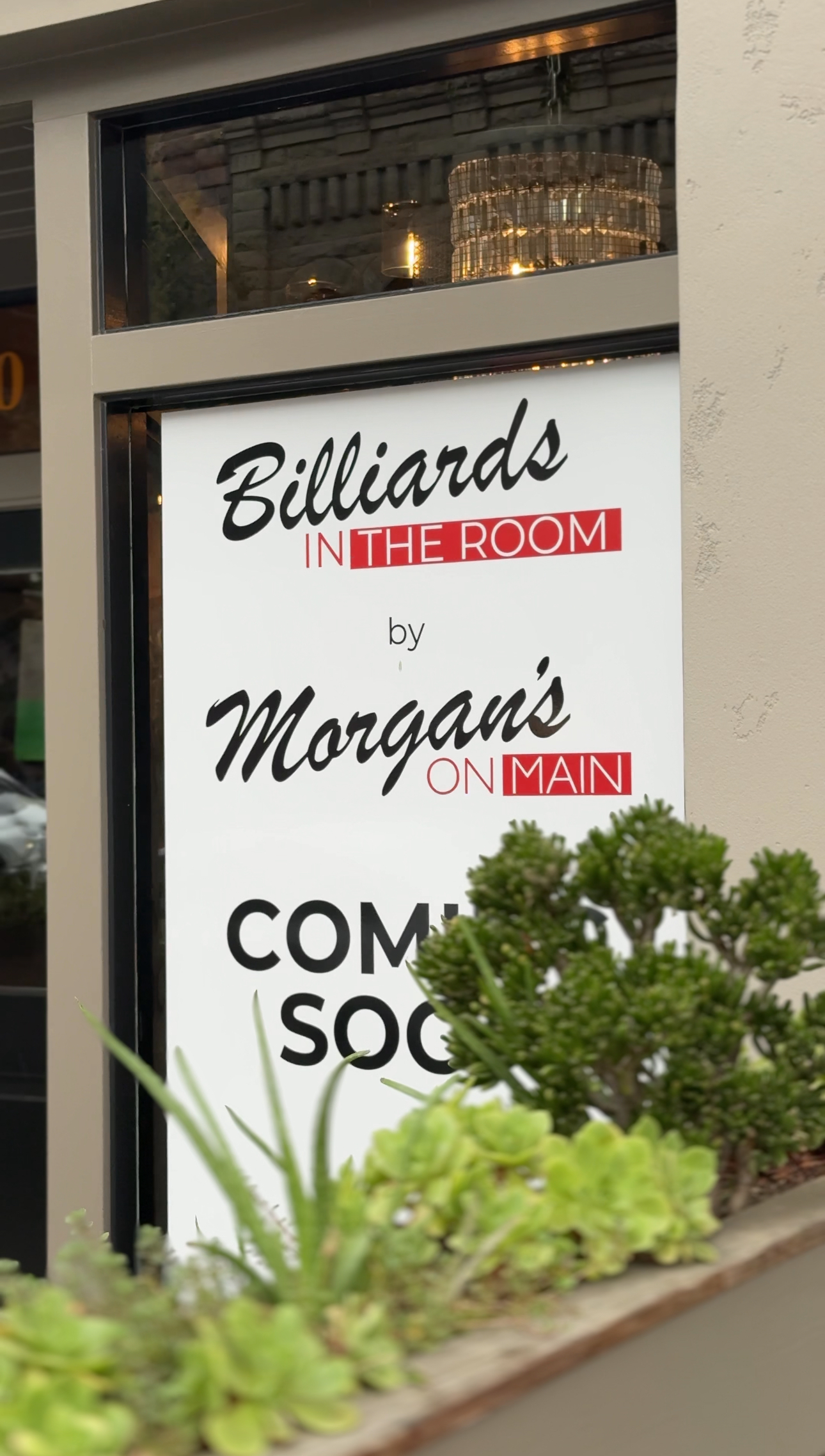 Sign: "Billiards in the Room by Morgan's on Main" "Coming Soon". Beige building, plants in foreground.