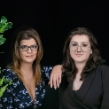 Two women with glasses posing in front of a black background.
