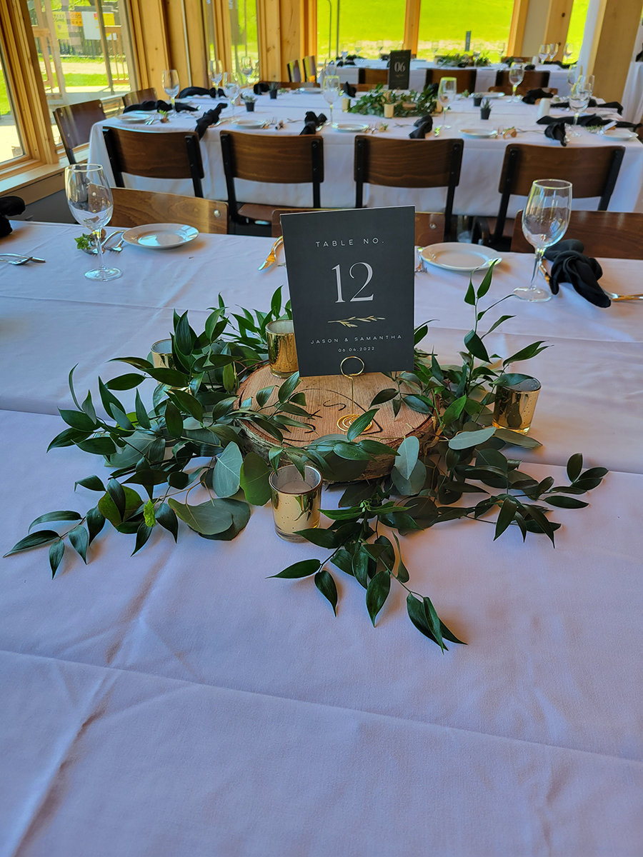 A table centerpiece at a wedding reception: a number 12 sign, greenery, gold candle holders, on a white tablecloth.