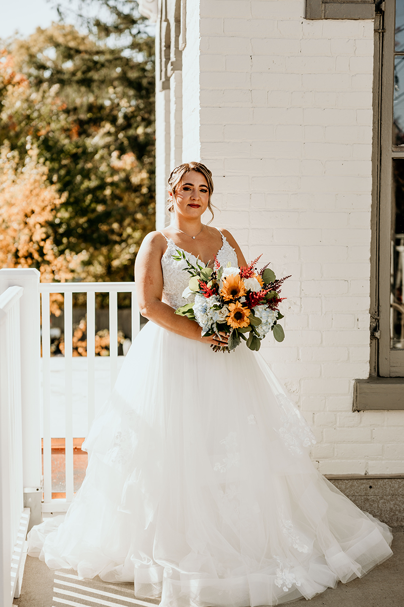 Bride in a white wedding dress, holding a bouquet, standing on a porch in front of a white brick wall.