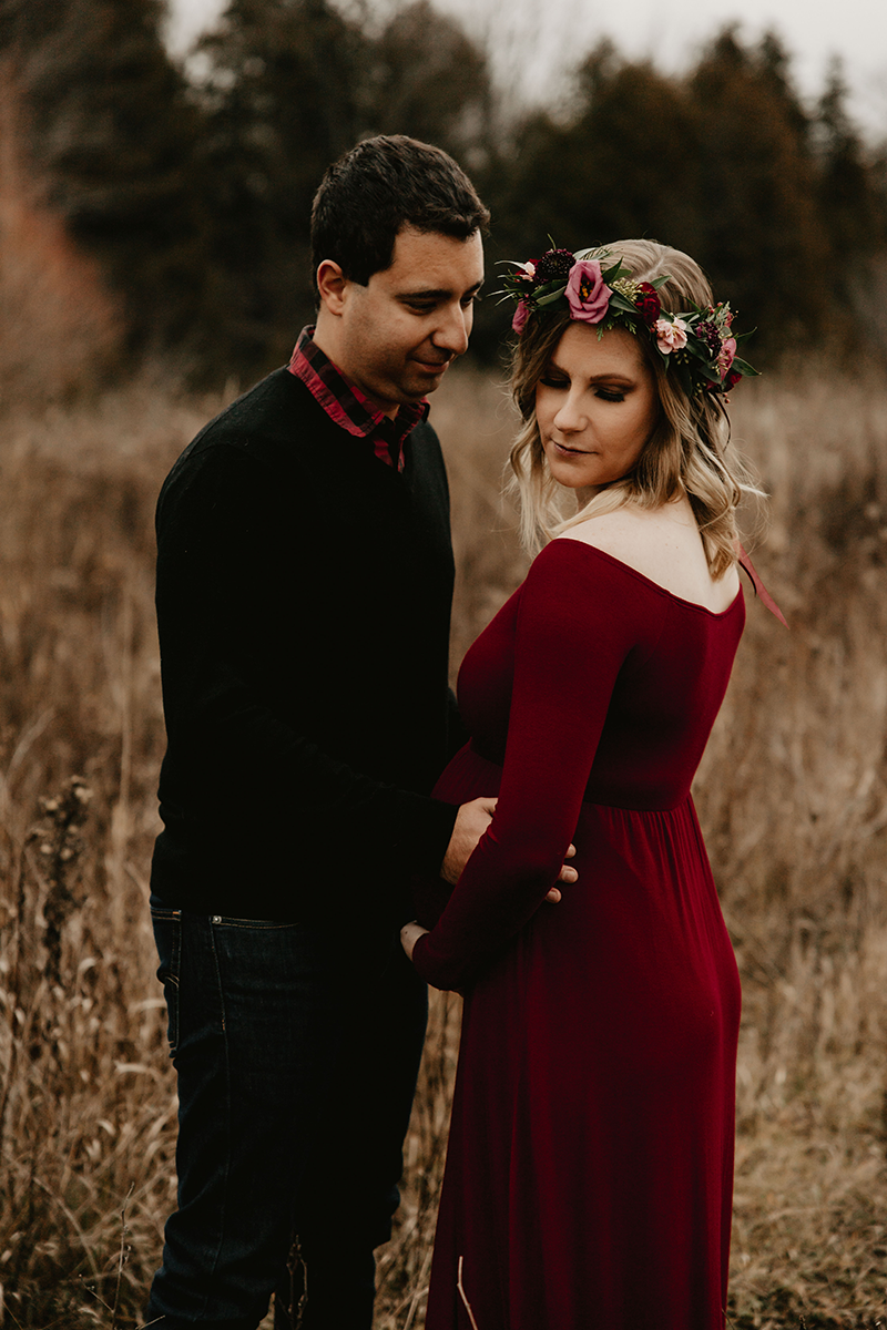 A pregnant woman in a burgundy dress and a flower crown stands with a man in a field. The man looks at her lovingly.