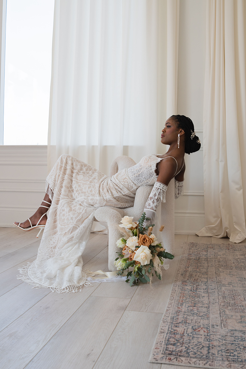Bride in a lace wedding dress reclines on a white bench, holding a bouquet. She poses in a bright room with neutral tones.