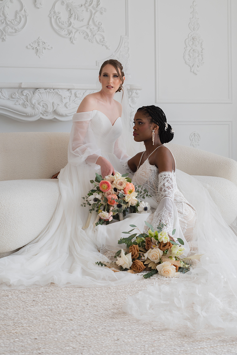 Two women in wedding dresses, one fair-skinned and one black, sit together on a cream-colored sofa, holding bouquets.