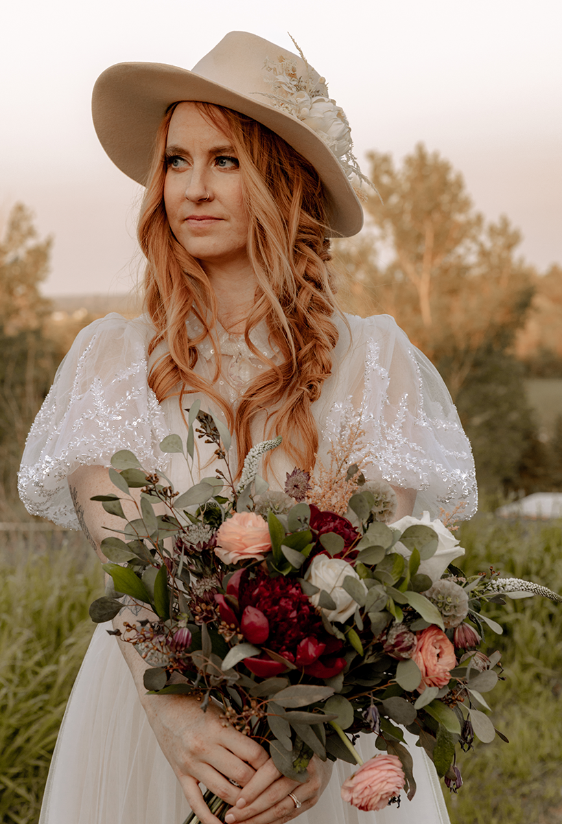 A red-haired woman in a cowboy hat and sparkly dress holds a bouquet. She gazes off to the side in an outdoor setting.