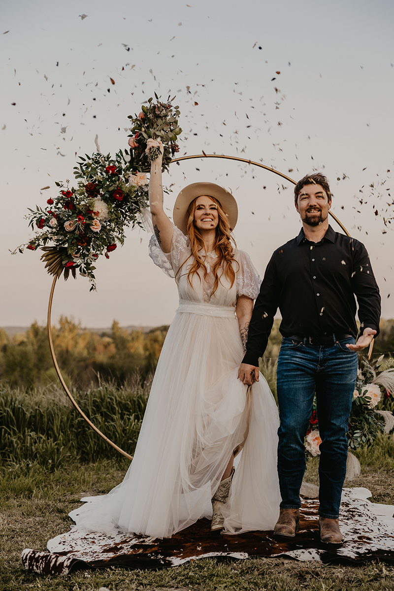 Newlyweds celebrate their wedding outdoors. The bride, in a white dress and hat, raises her bouquet as confetti falls.