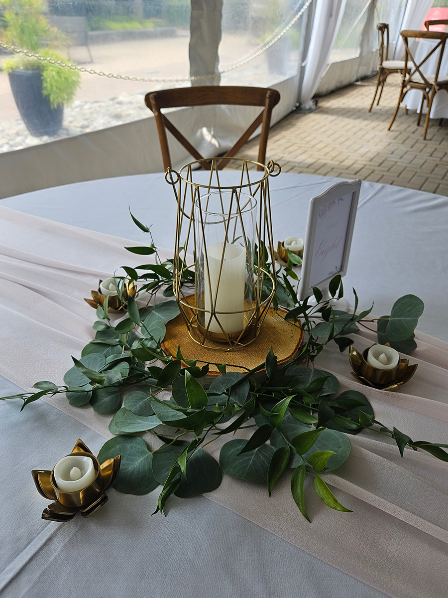 Wedding table centerpiece with a gold lantern, greenery, candles, and a wooden base on a white tablecloth.