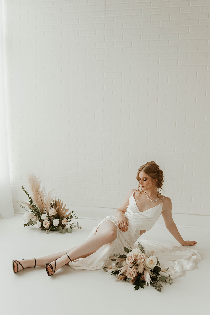 Woman in white gown reclines on white floor with flowers, a bouquet, and a floral arrangement.