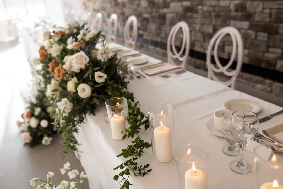 Long white wedding reception table decorated with floral arrangements and candles. Elegant place settings are arranged.