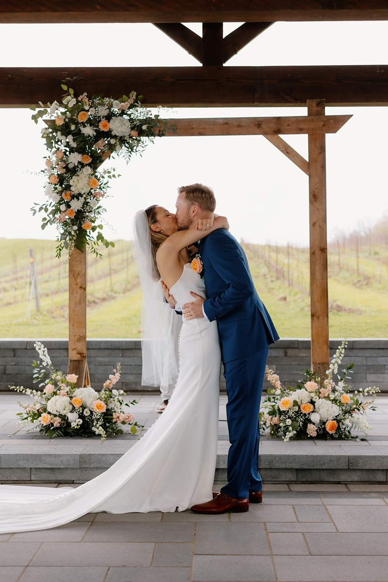 Newly married couple kissing under a floral-decorated wooden arch. The bride wears a white dress and veil, the groom a suit.