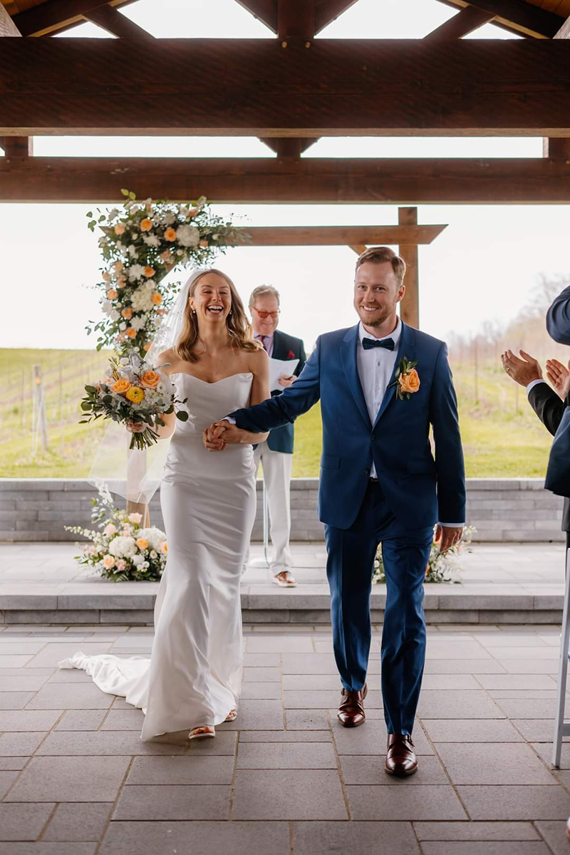 Newly married couple exits ceremony under a covered patio. The bride wears a white dress, the groom a blue suit.