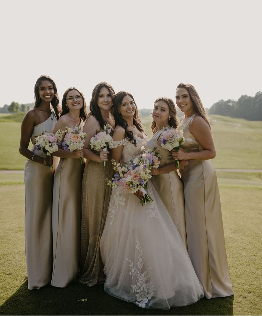 A bride in a white dress poses with her bridesmaids in gold dresses, holding bouquets, on a golf course.