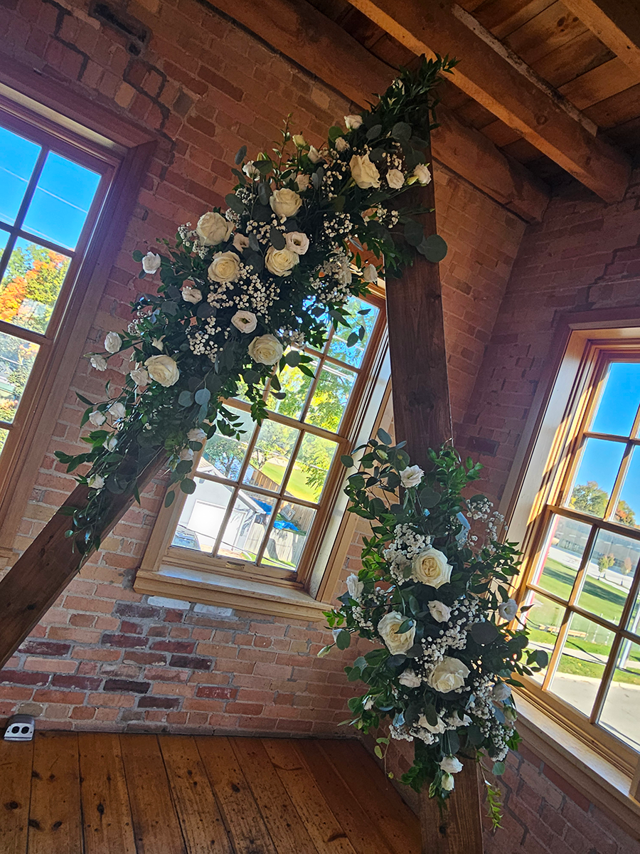 Floral archway with white roses and greenery, set against brick and wooden beams, framing a bright window view.