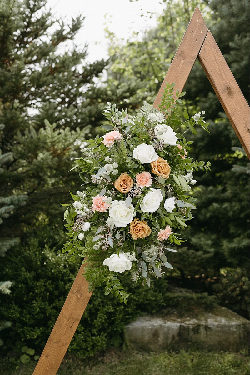 Wooden A-frame arch decorated with white, peach, and tan flowers and greenery, set in a garden.