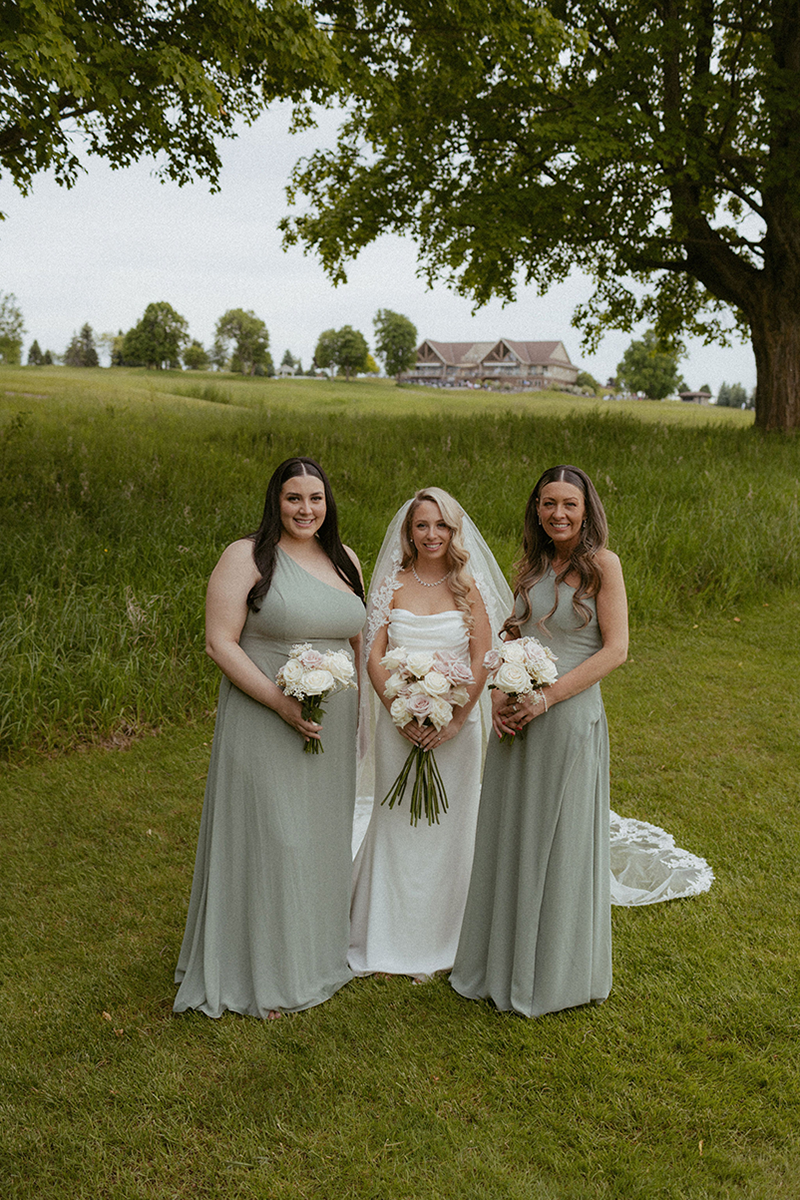 Bride in white dress with two bridesmaids in sage green dresses, posing in a grassy field.