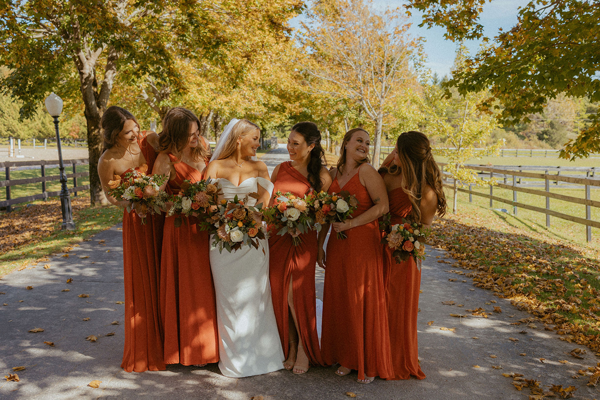 Bride and bridesmaids in rust-colored dresses stand on a path, holding bouquets.