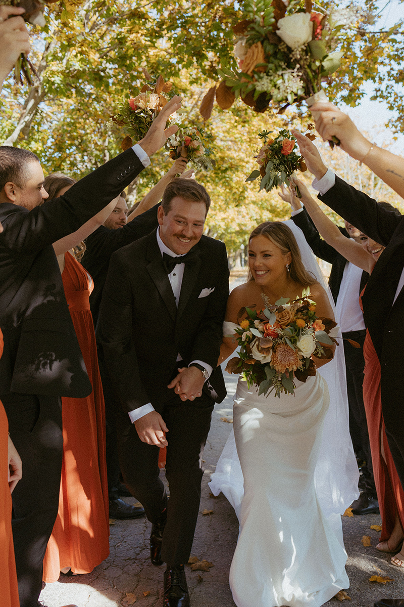 Newly married couple walking through a flower archway created by their wedding party, smiling.