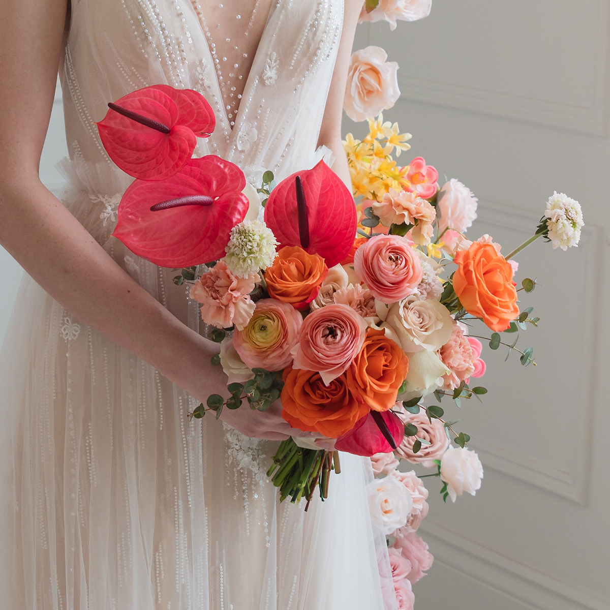 Bride in a white gown holding a vibrant bouquet with orange and pink flowers, including anthuriums, in front of a white wall.