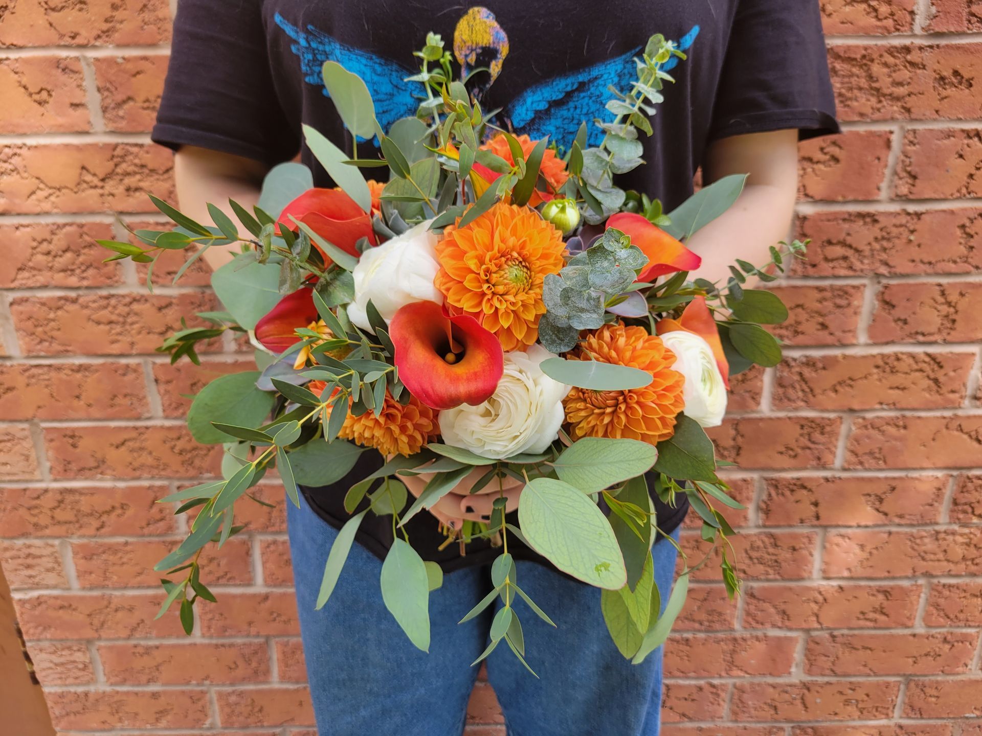 Person holding a bouquet of orange and white flowers, with green leaves, in front of a brick wall.
