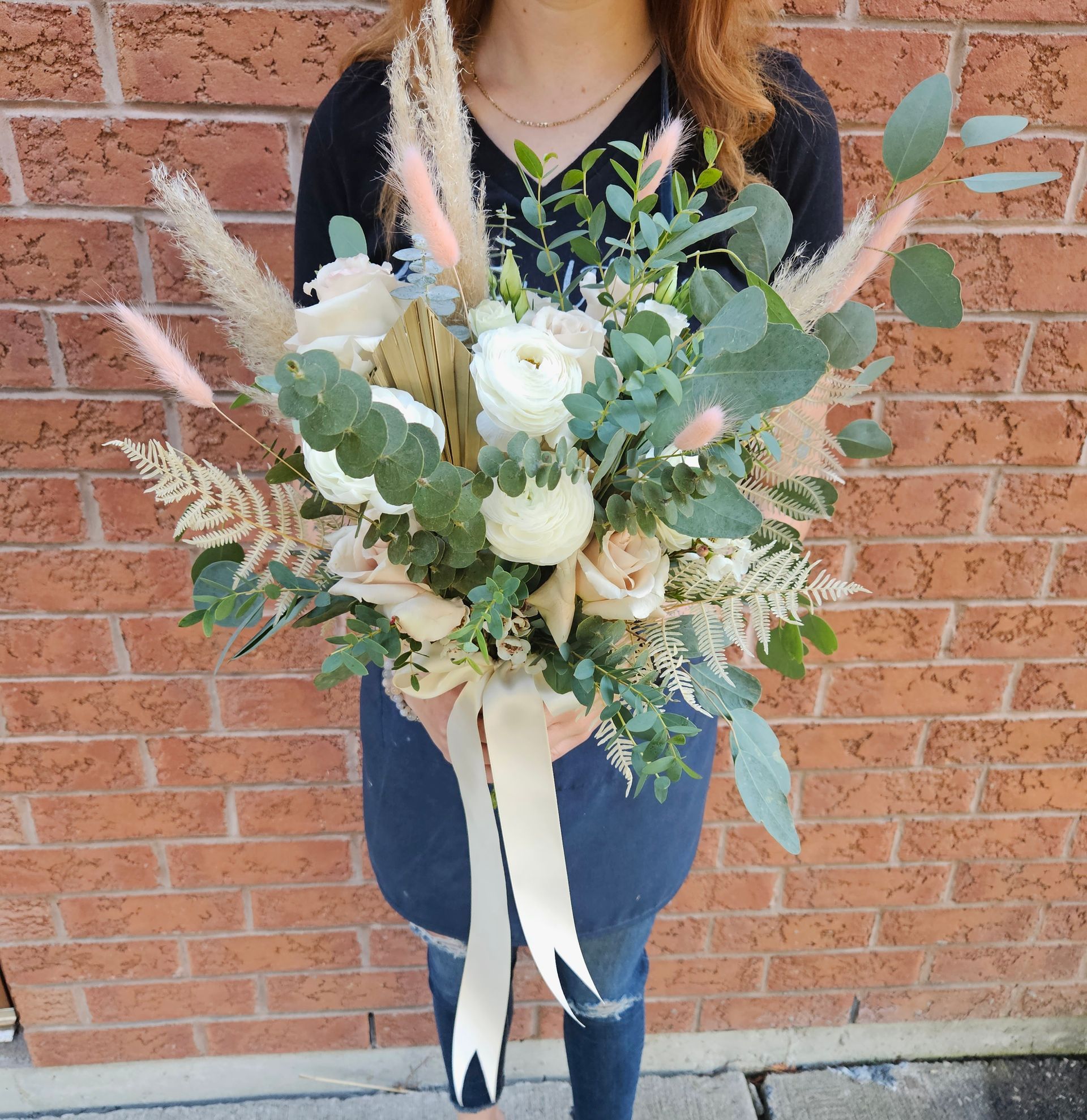 Woman holding a bouquet of cream-colored flowers and greenery against a brick wall.