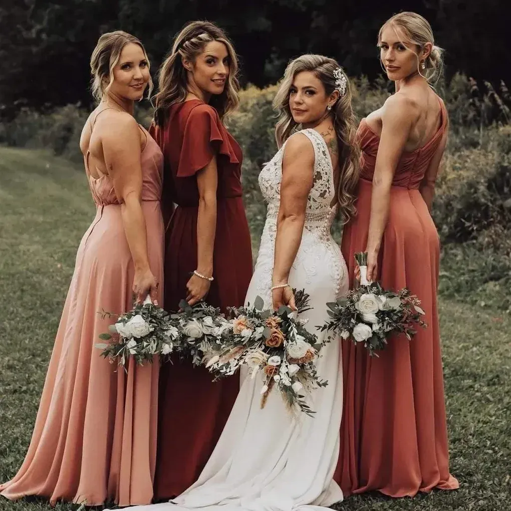 Bride and bridesmaids in a field. Bride in white lace, bridesmaids in varying shades of red dresses holding bouquets.