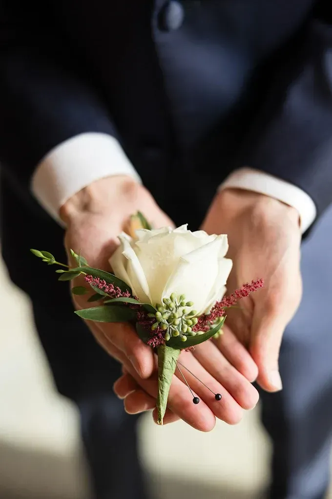 A person in a navy suit holds a white rose boutonniere with greenery in their hands.