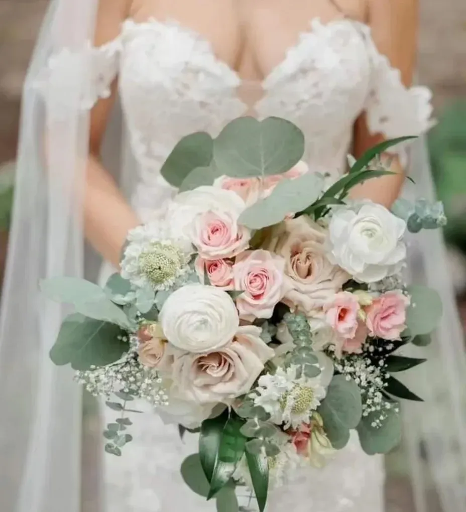 Bride holding a bouquet of light pink and cream roses, eucalyptus, and baby's breath, wearing a white lace wedding dress.