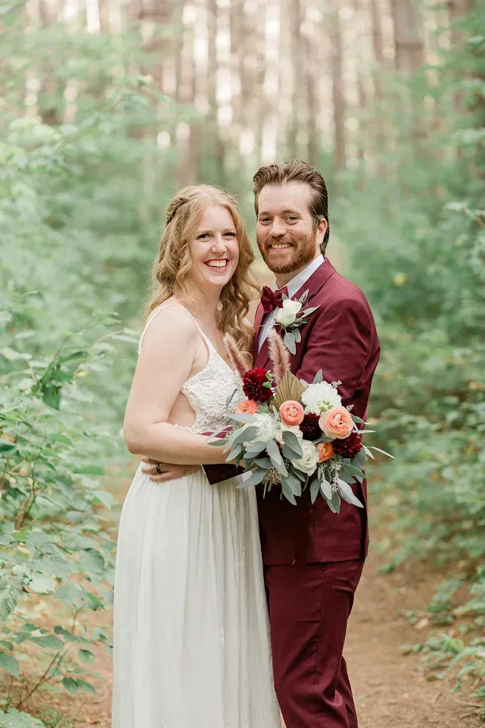 Newlyweds smiling in a forest setting. Bride in white dress, groom in a maroon suit, holding a bouquet.