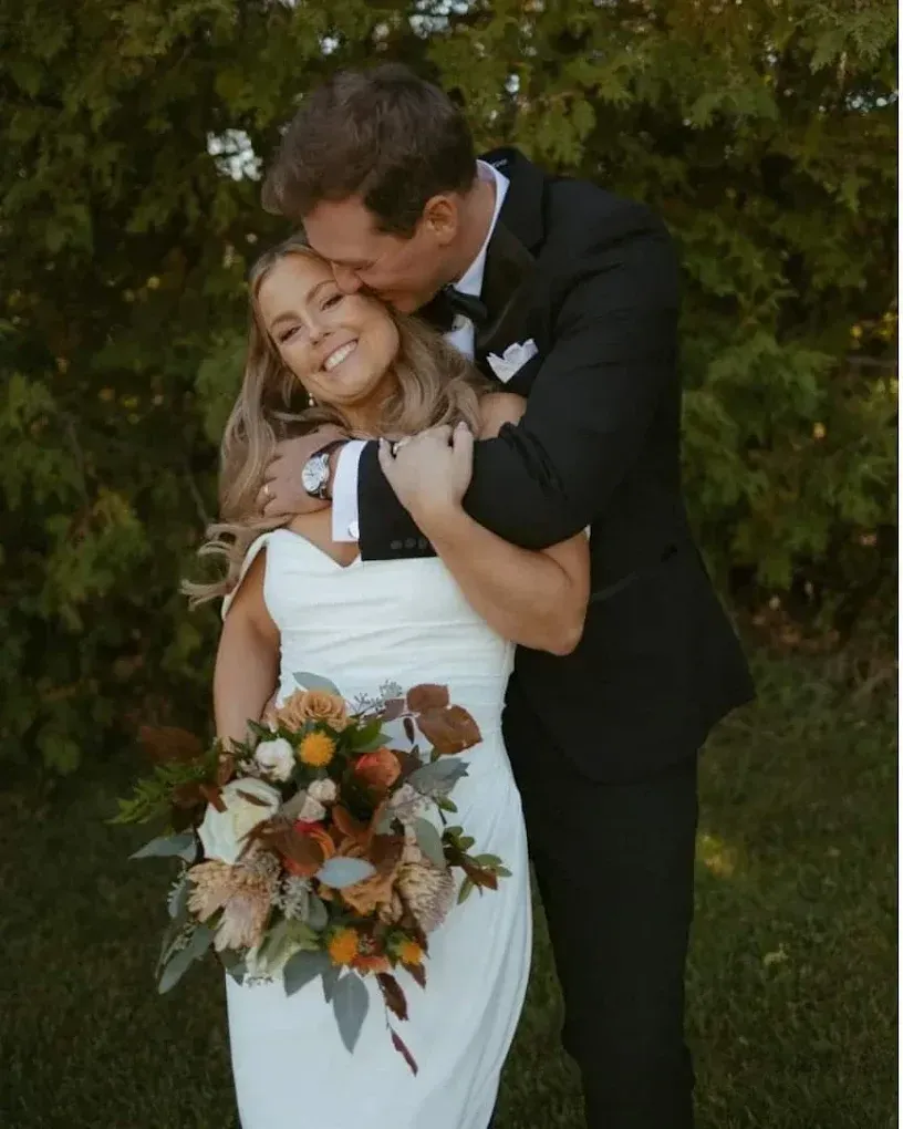 A smiling bride in a white gown is embraced and kissed on the cheek by her groom.
