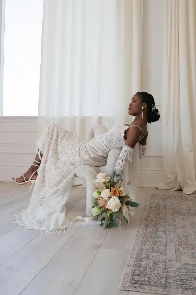 Bride in a lace wedding dress reclines on a white chair, holding a bouquet. She wears gloves and earrings.