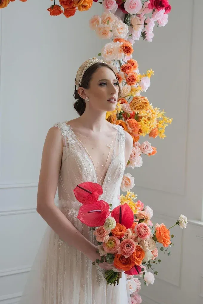 Bride in a white gown holding a colorful bouquet, standing before a floral arch. Her hair is up and she looks to the side.