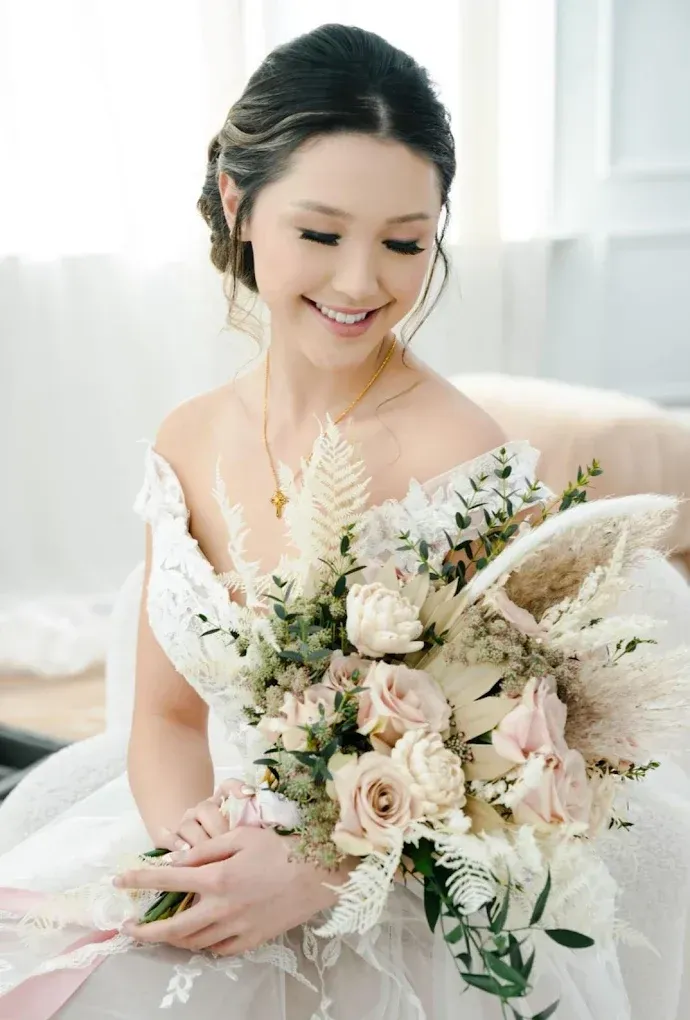 Bride in a white off-the-shoulder gown smiles, holding a bouquet with soft pink and cream flowers and greenery.