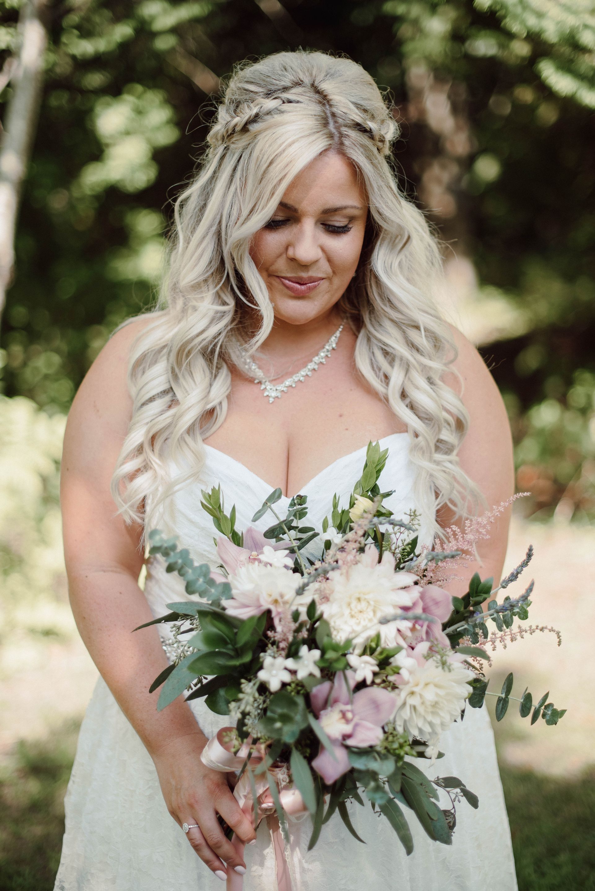 Bride in a white dress holds a bouquet of flowers, looking down. She has blonde hair in a half-up, half-down style.