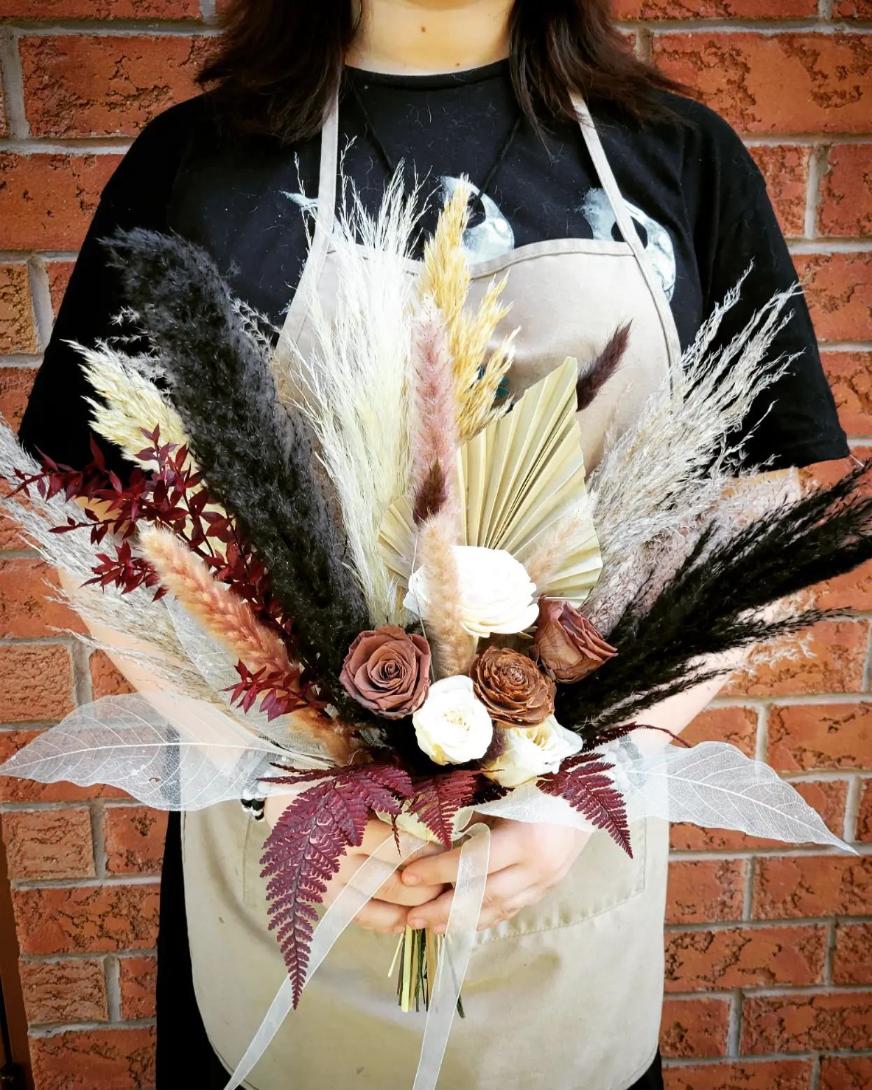 Woman holding a bouquet of dried flowers with beige and black accents, tied with a ribbon; brick wall background.