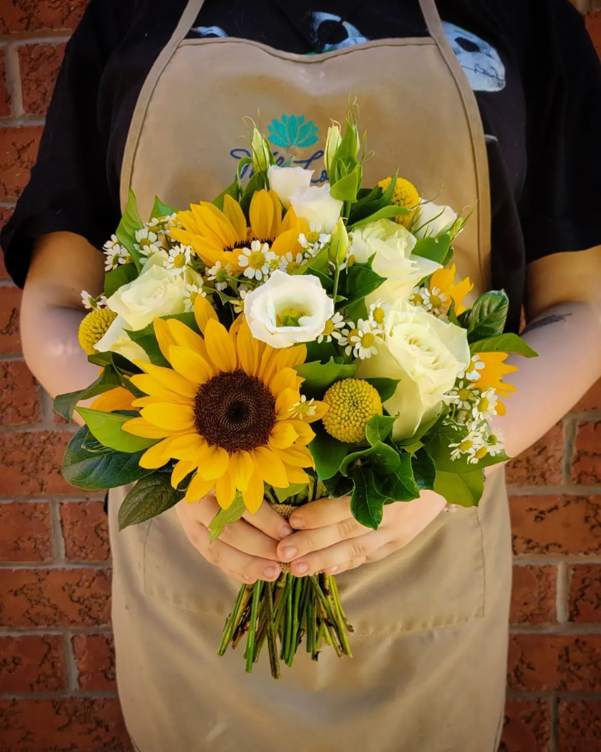 Person in apron holding a bouquet with sunflowers, white roses, and daisies in front of a brick wall.
