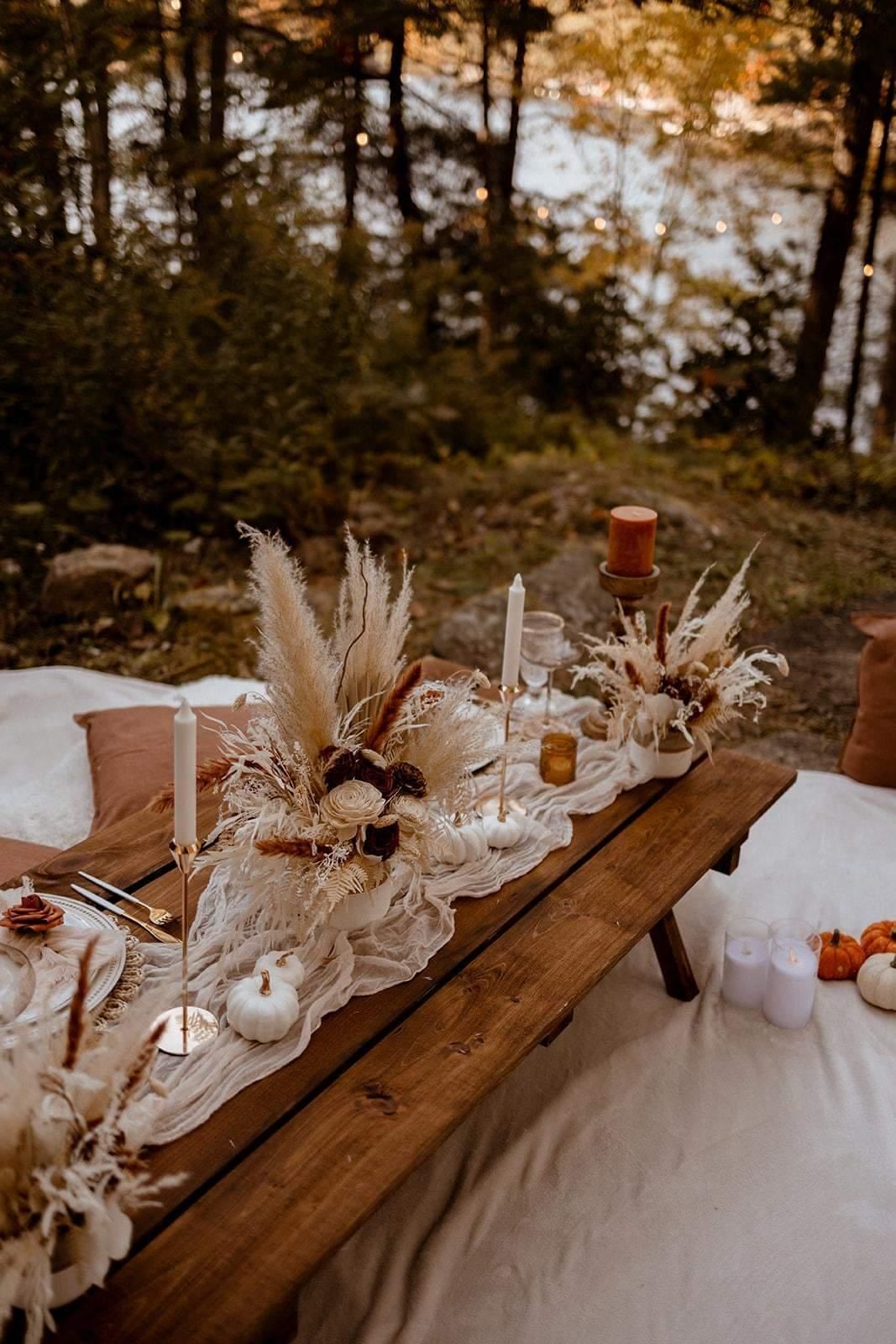 Wooden picnic table set for an outdoor fall celebration with floral centerpieces, candles, and pumpkins.