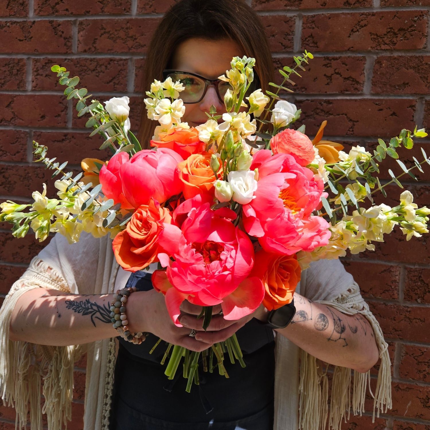 Woman holding a vibrant bouquet of orange and pink flowers in front of a brick wall.