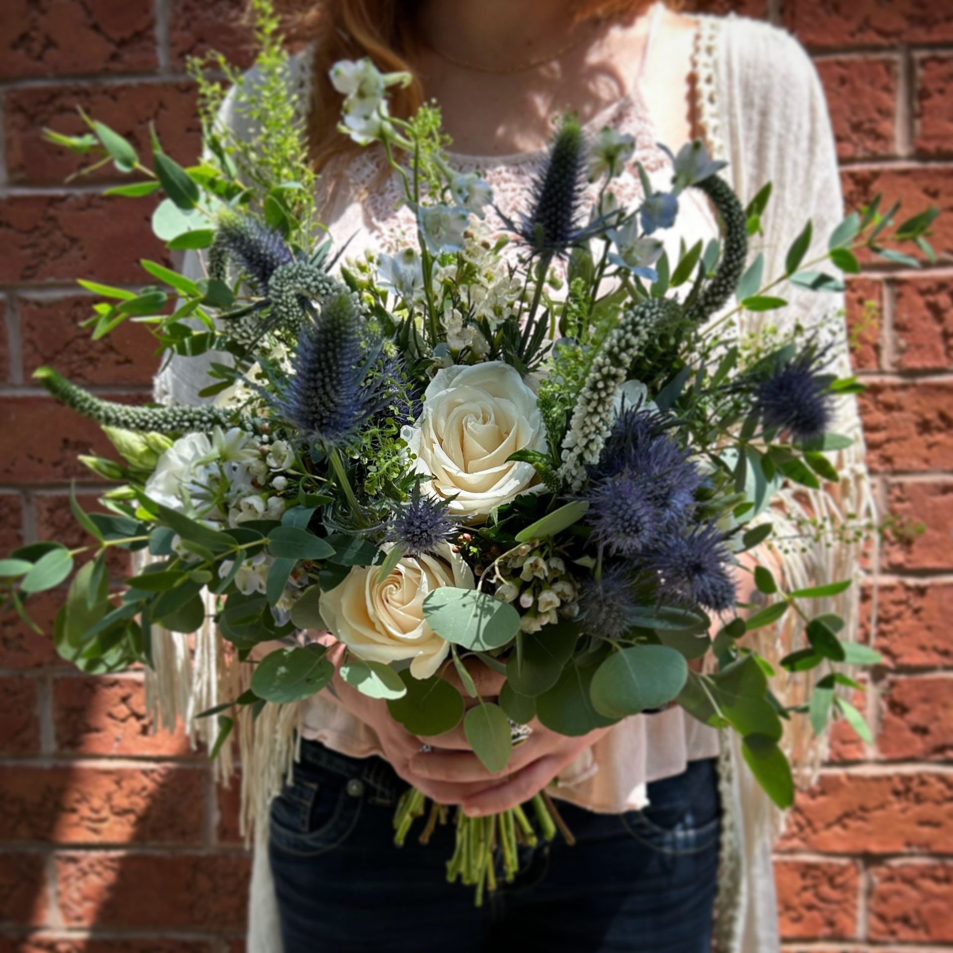 Woman holding a bouquet of white roses, blue thistle, and greenery. Against a brick wall.