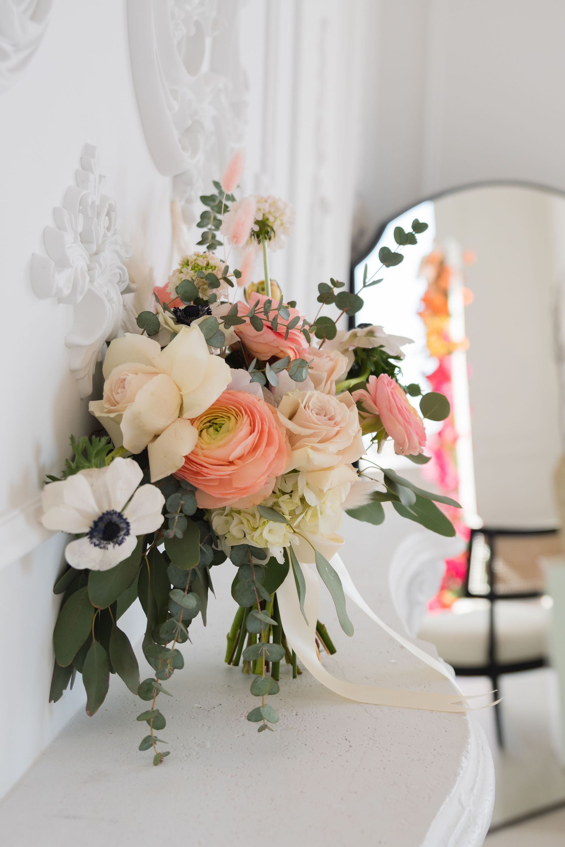 Bridal bouquet with peach and white flowers, greenery, and a cream ribbon, resting on a white decorative mantel.