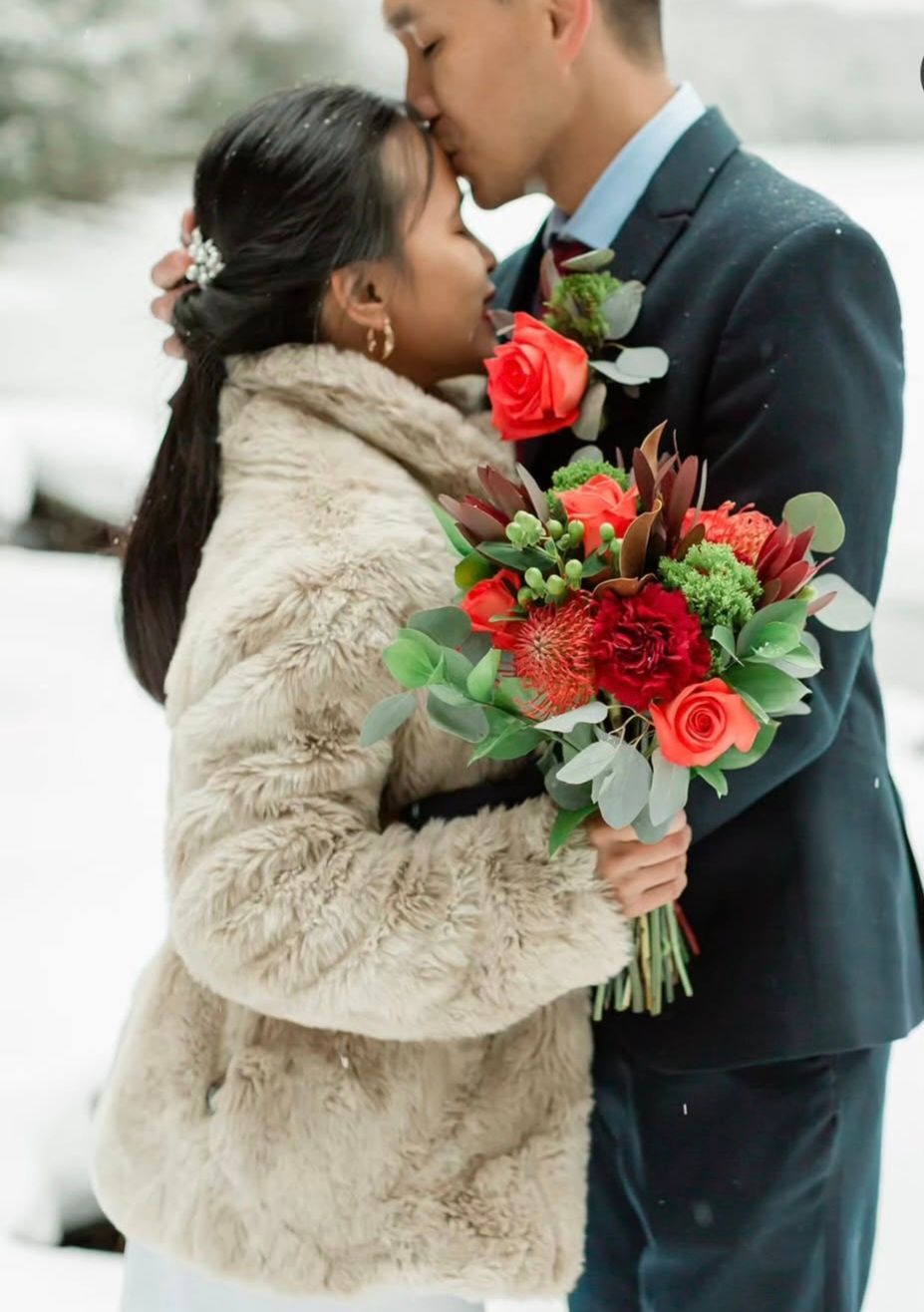 Couple embracing, holding bouquet outdoors in snow. Man kissing woman’s forehead; she smiles. Beige faux fur jacket.