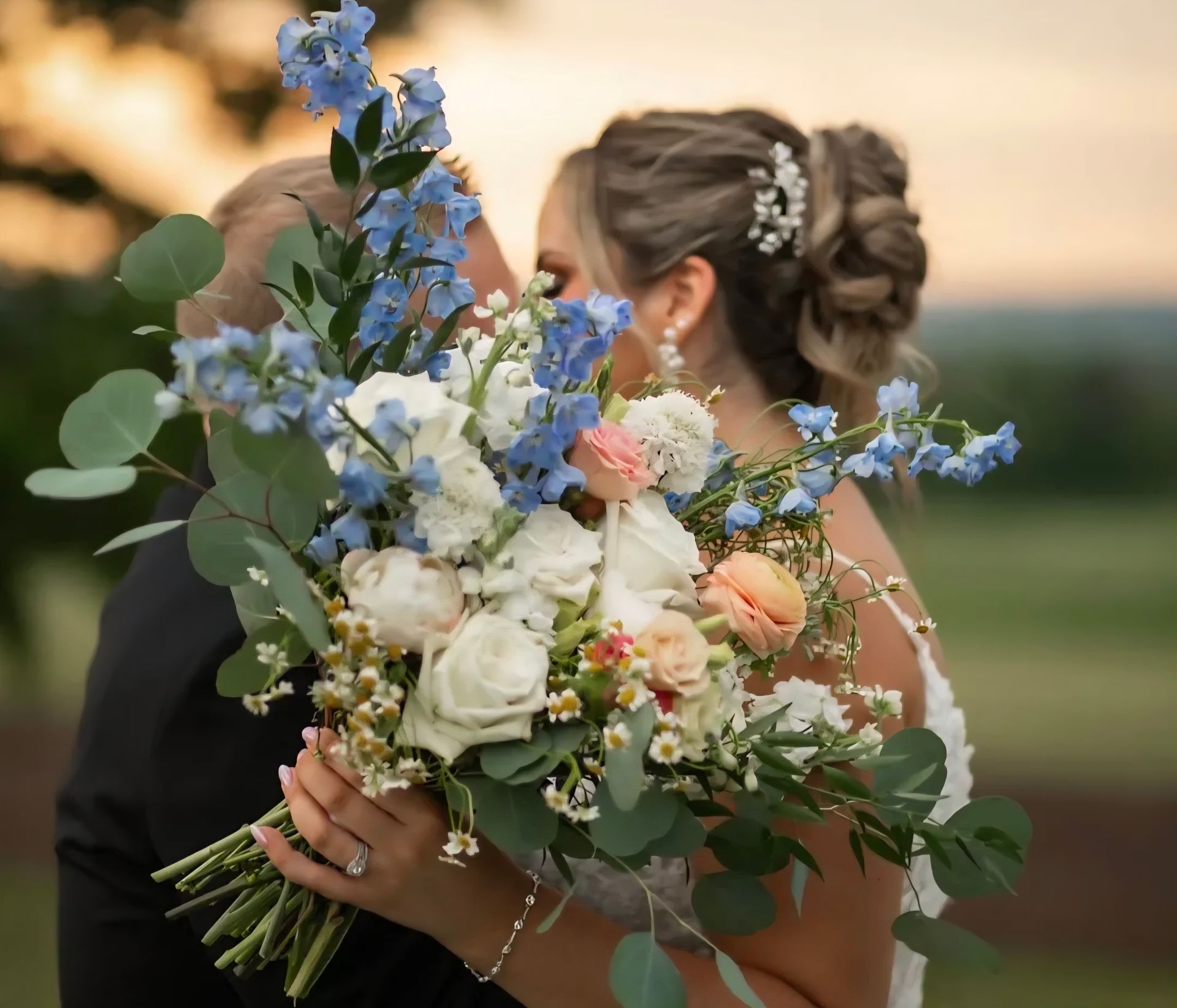 Bride and groom kissing, holding a bouquet of blue, white, and peach flowers, with a sunset in the background.