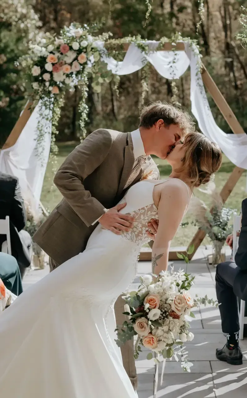 Couple kissing, bride in white dress, groom in brown suit, at outdoor wedding ceremony.