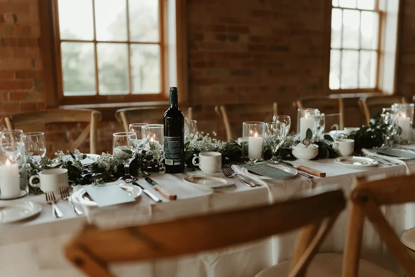 Long dining table set for an event, with greenery, candles, and wine bottles. Windows with natural light in the background.